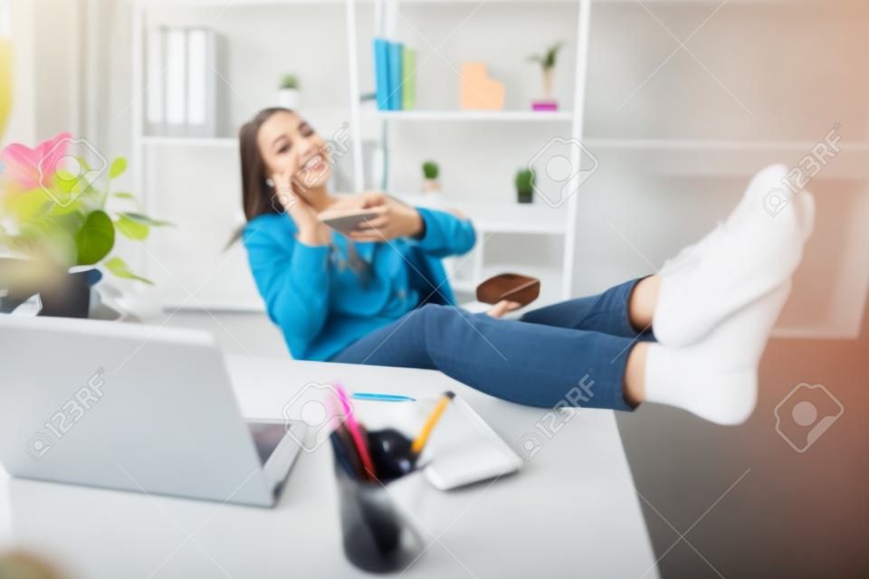 Portret uroczej stylowej dziewczyny kładącej nogi na stole, mając przerwę w wolnym czasie za pomocą smartfona rozmawia z przyjacielem ciesząc się odpoczynkiem, bawiąc się na stanowisku pracy