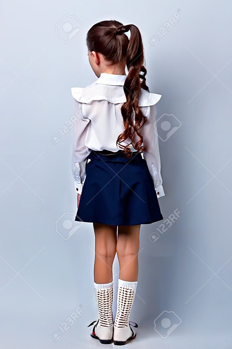 Schnappschuss-Rückansicht des netten entzückenden stilvollen Mädchens mit den lockigen Zöpfen im formellen weißen Blusenhemd, im kurzen blauen Rock, in den Gamaschen, in den Schuhen. Isoliert über grauem Hintergrund