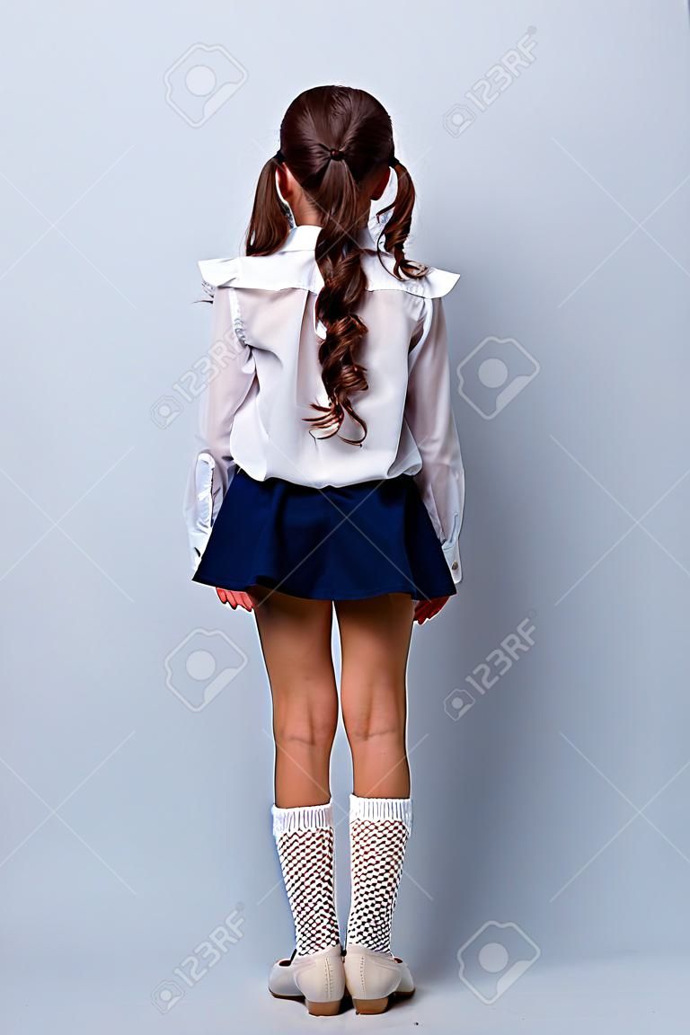 Snap shot rear back view of nice adorable stylish girl with curly pigtails in formal white blouse shirt, short blue skirt, gaiters, shoes. Isolated over grey background