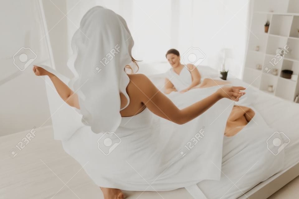 A woman in white towel flashing her body to her husband lying on the bed. rear view