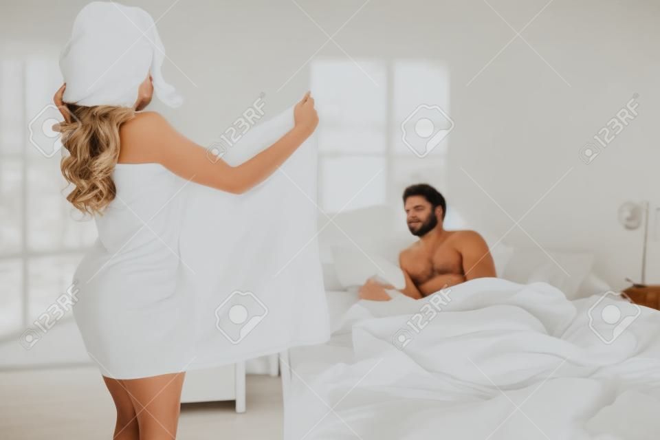 A woman in white towel flashing her body to her husband lying on the bed. side view