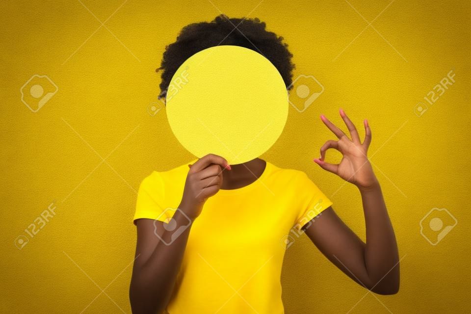 Close up photo beautiful amazing she her dark skin lady hiding face okey symbol fingers round circle paper unrecognized opinion wear casual white t-shirt isolated yellow bright vibrant background