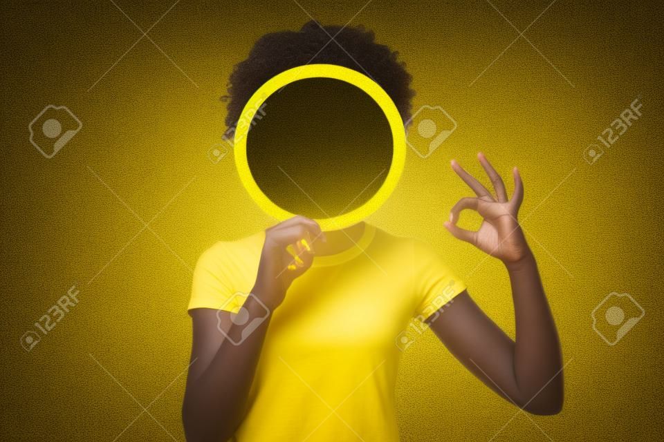 Close up photo beautiful amazing she her dark skin lady hiding face okey symbol fingers round circle paper unrecognized opinion wear casual white t-shirt isolated yellow bright vibrant background