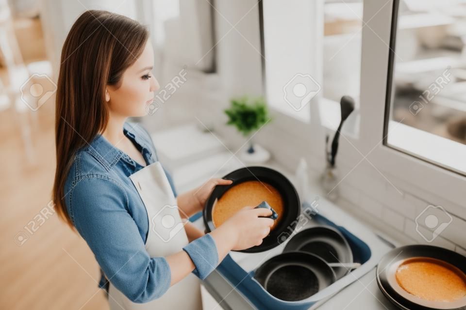 Close up side profile photo attentive beautiful she her lady cleaner bright kitchen hold dirty plate spring cleanup preparing meeting holiday gathering wear casual jeans denim shirt apron flat indoors