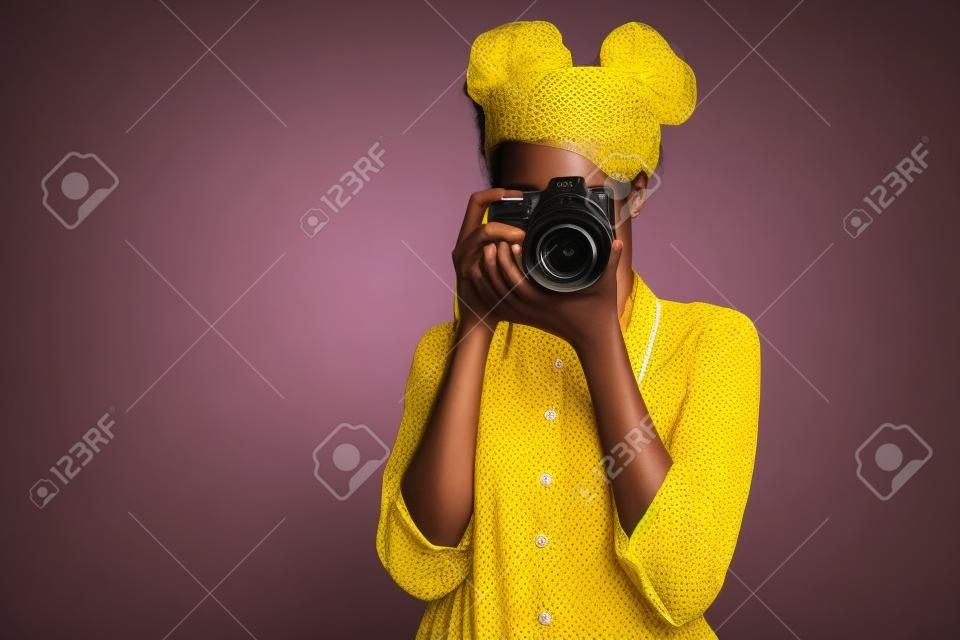 Foto di una straordinaria signora dalla pelle scura che tiene una foto digicam nelle mani che fotografa, visite turistiche straniere all'estero indossano pantaloni della camicia gialla isolati sullo sfondo di colore viola