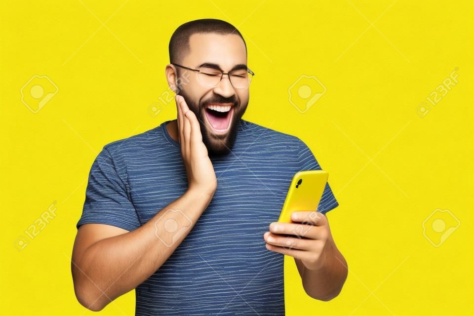 Photo of ecstatic overjoyed man browsing through his telephone rejoicing in receiving, positive notifications in t-shirt isolated vivid color yellow background