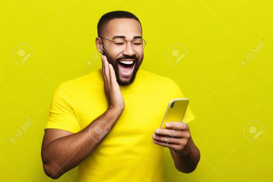 Foto de hombre lleno de alegría extático navegando a través de su teléfono regocijándose en recibir, notificaciones positivas en camiseta aislado fondo amarillo de colores vivos
