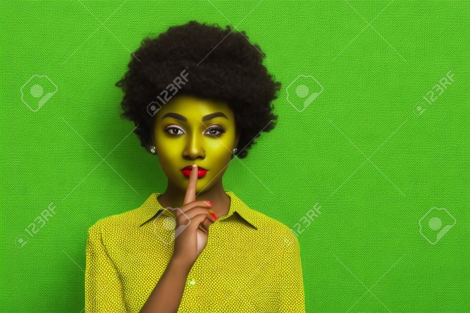 Photo of pretty dark skin wavy lady holding finger on lips asking keep silence looking wondered empty space wear trendy green dotted shirt blouse isolated yellow color background