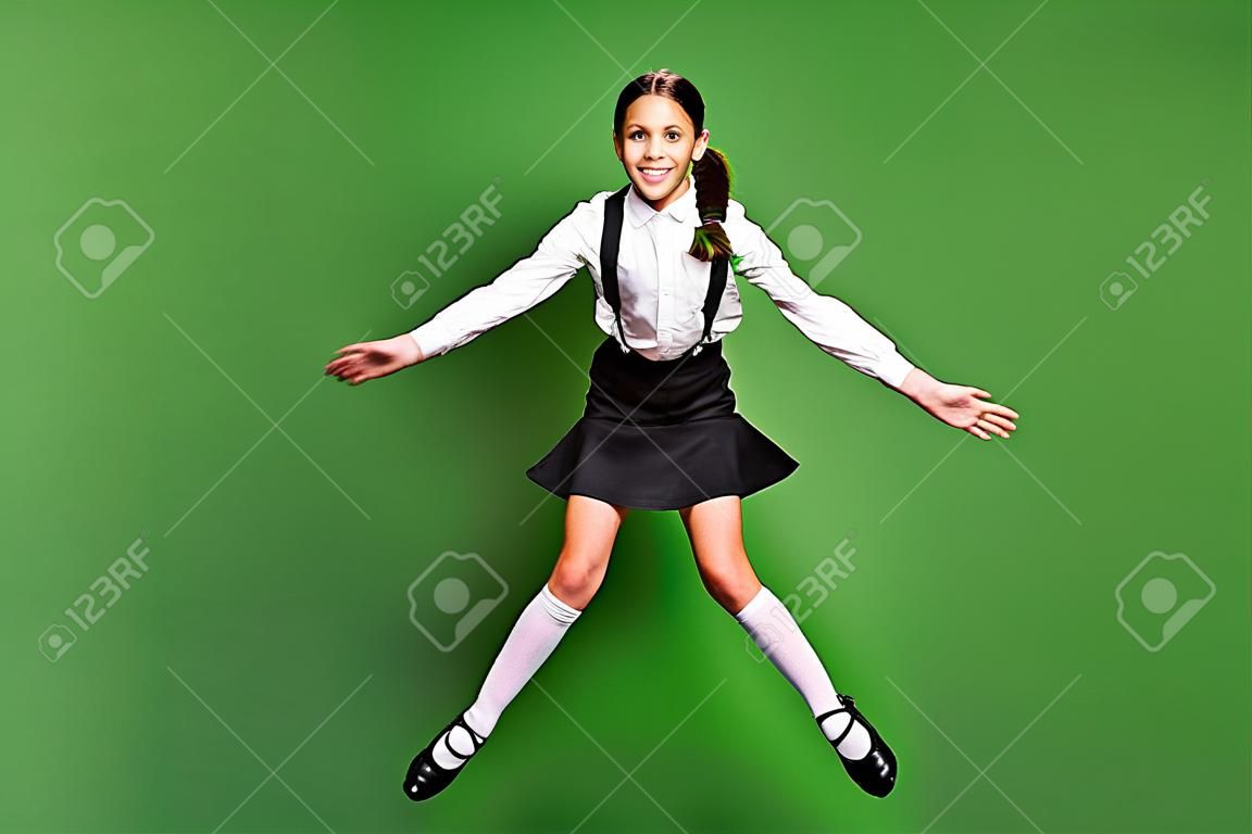 Retrato fotográfico de una colegiala morena con coletas saltando con las piernas abiertas y las manos juntas aisladas en un fondo de color verde vivo