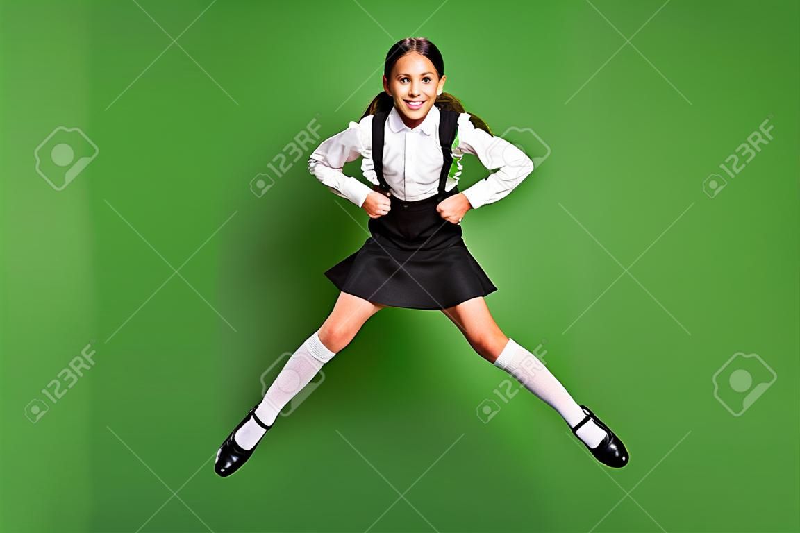 Retrato fotográfico de una colegiala morena con coletas saltando con las piernas abiertas y las manos juntas aisladas en un fondo de color verde vivo