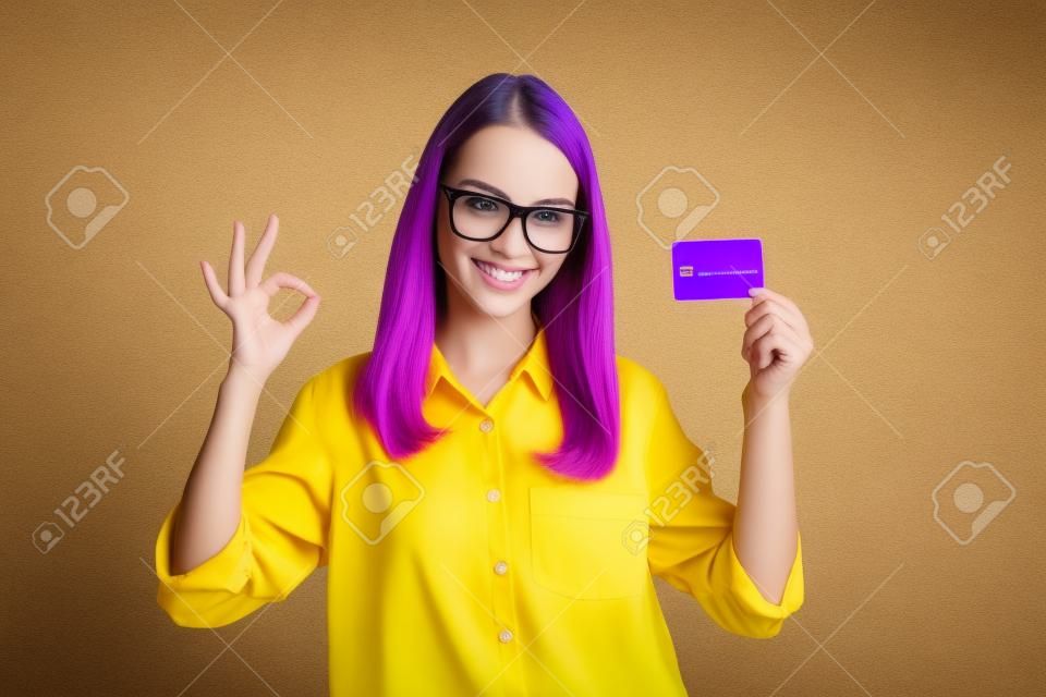 Photo of young lady raise hand show okey hold debit card toothy smile wear spectacles yellow shirt isolated purple color background