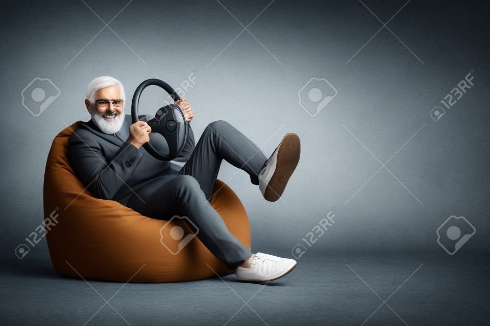 Portrait of bearded grey-haired man sitting in bag chair having fun holding steering wheel