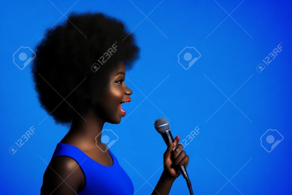 明るい青色の背景で隔離のコピースペースを見てマイクで歌っているかなり黒い肌の女の子の写真の横顔の肖像画