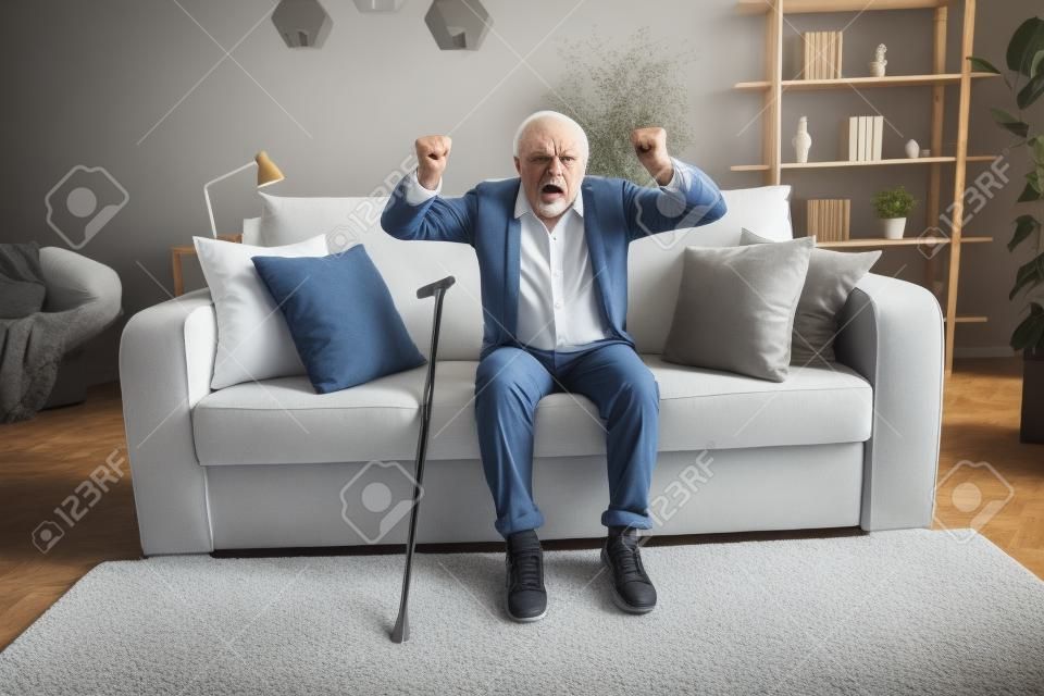 Foto de un anciano enojado vestido con camisa blanca sentado en un sofá levantando los puños en el interior de una casa plana