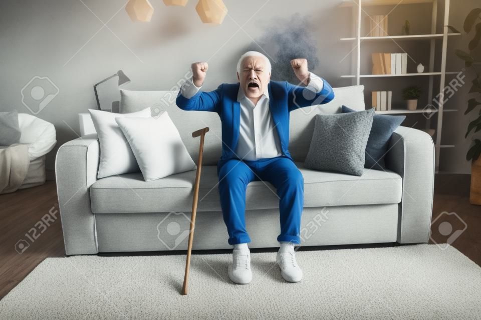 Foto de un anciano enojado vestido con camisa blanca sentado en un sofá levantando los puños en el interior de una casa plana