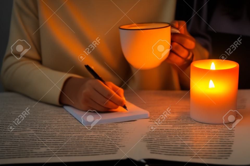 female hands with pen and cup of tea or coffee writing on notebook in candle light