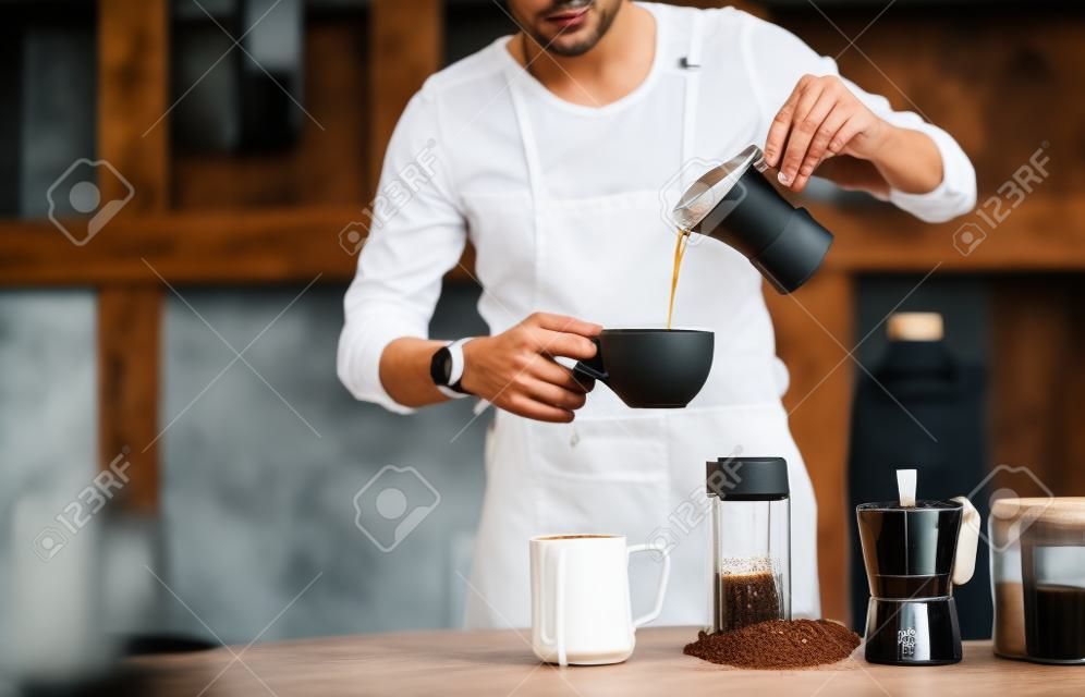 Barista gießt Wasser in Moka-Kaffee in eine Tasse mit Flasche Milch und Brauausrüstung auf einem Holztisch.