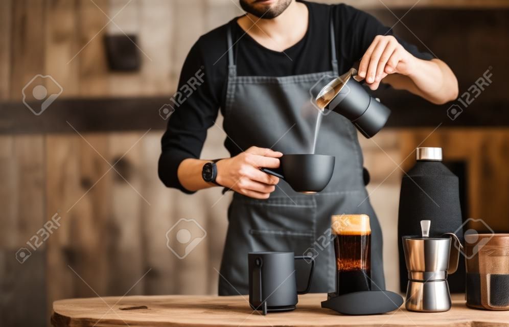 Barista gießt Wasser in Moka-Kaffee in eine Tasse mit Flasche Milch und Brauausrüstung auf einem Holztisch.