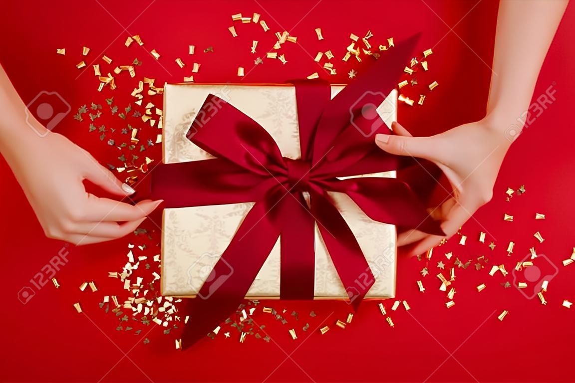 Female hands tying a bow on gift box with a red ribbon on wine background. Concept of a gift for the holidays, birthday, christmas Flat lay, top view