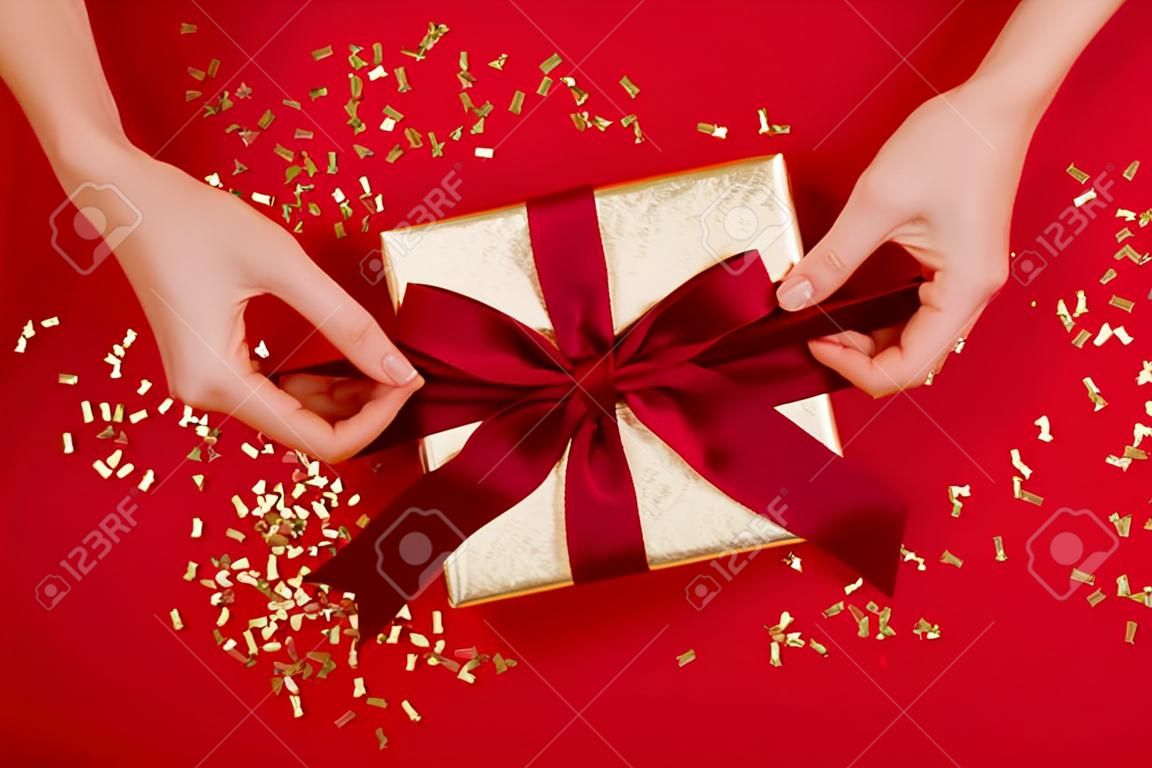 Female hands tying a bow on gift box with a red ribbon on wine background. Concept of a gift for the holidays, birthday, christmas Flat lay, top view