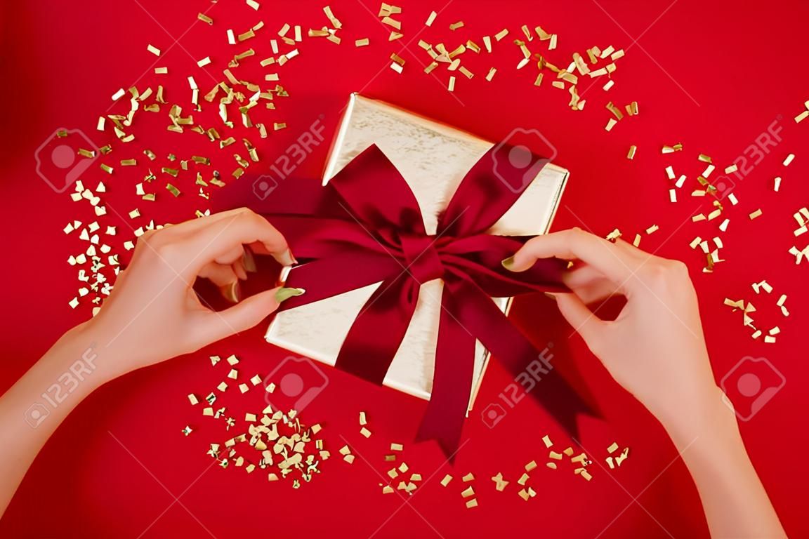 Kobiece dłonie zawiązujące kokardkę na pudełku z czerwoną wstążką na tle wina Koncepcja prezentu na święta Urodziny Boże Narodzenie Mieszkanie leżało Widok z góry
