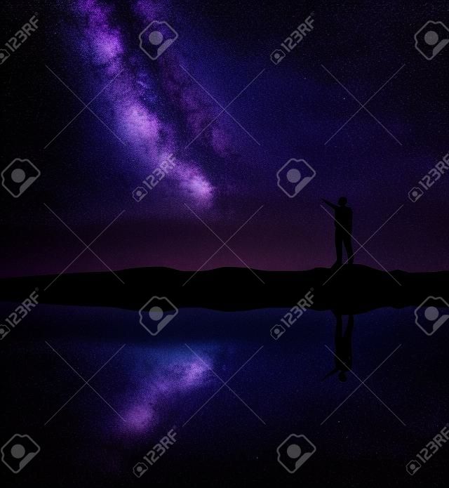 Milky Way. Silhouette of a standing man pointing finger in night starry sky on the mountain near the river with sky reflection in water. Night landscape with galaxy. Purple milky way and man. Universe