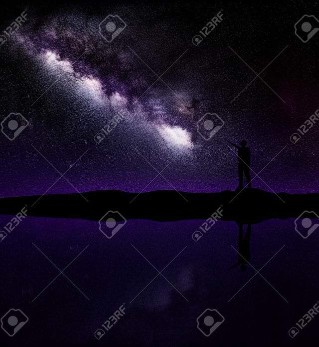 Milky Way. Silhouette of a standing man pointing finger in night starry sky on the mountain near the river with sky reflection in water. Night landscape with galaxy. Purple milky way and man. Universe