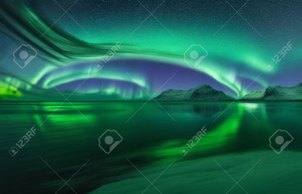 极光。北极光在罗弗敦群岛，挪威。与极光的满天星斗的蓝天。夜冬季景观与极光，海与天空反射，海滩，山脉，城市的灯光。绿色北极光