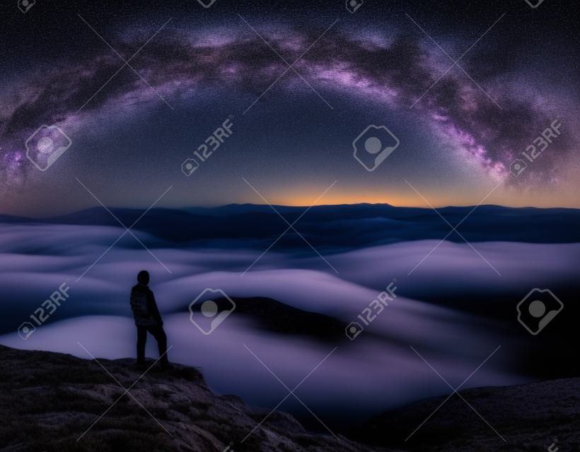 Man on the mountain peak and arched Milky Way over mountains in low clouds at night. Landscape with purple starry sky, Milky Way Arch, orange light, guy, hills in fog. Space and galaxy. Sky with stars