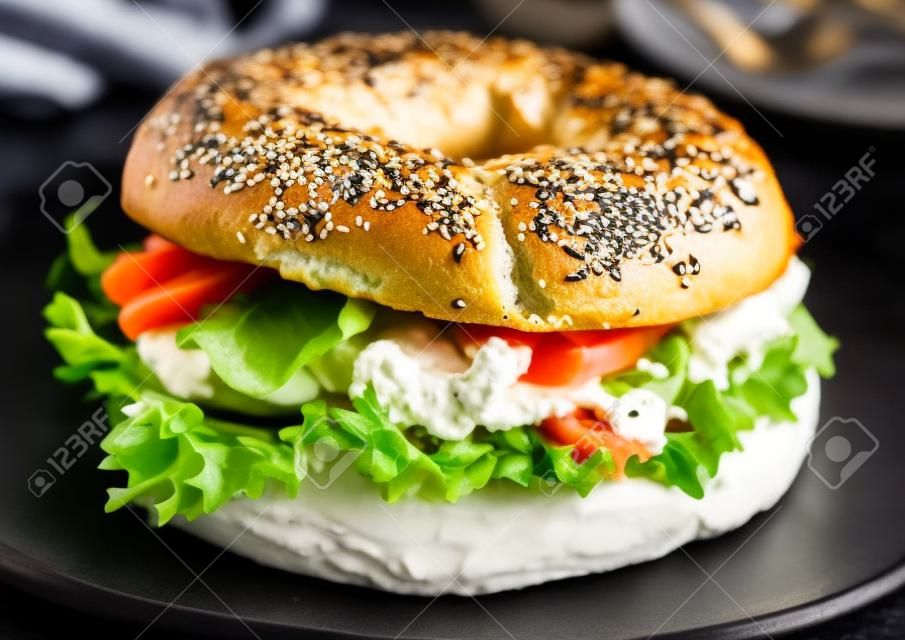 Fresh healthy bagel sandwich with salmon, ricotta and lettuce in black plate on dark kitchen table background.