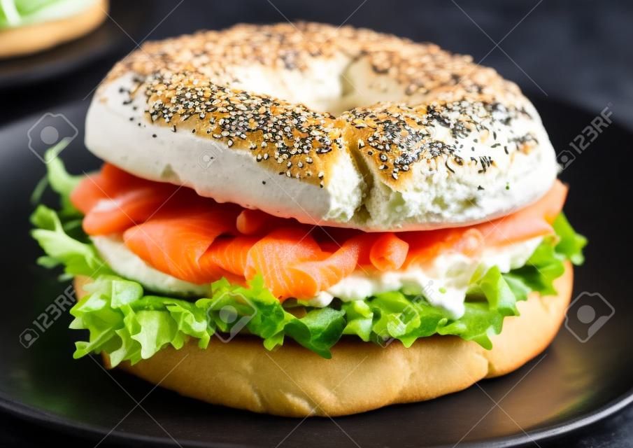 Verse gezonde bagel sandwich met zalm, ricotta en sla in zwarte plaat op donkere keukentafel achtergrond.
