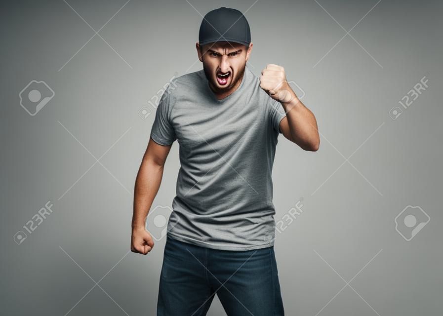 Angry Delivery man in gray t-shirt and cap threatens fist. Studio portrait of caucasian handsome guy in rage looking furious. Upset and mad courier isolated on white background.