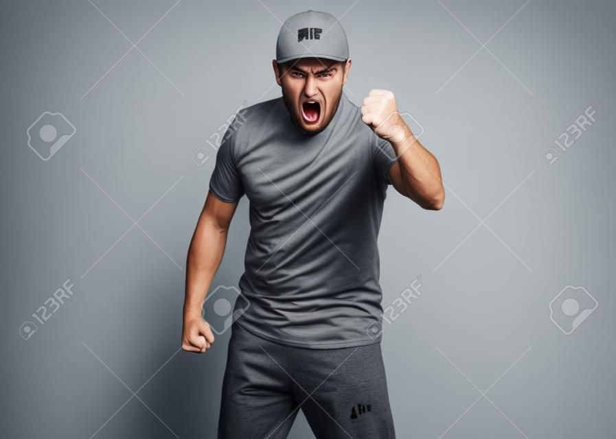 Angry Delivery man in gray t-shirt and cap threatens fist. Studio portrait of caucasian handsome guy in rage looking furious. Upset and mad courier isolated on white background.