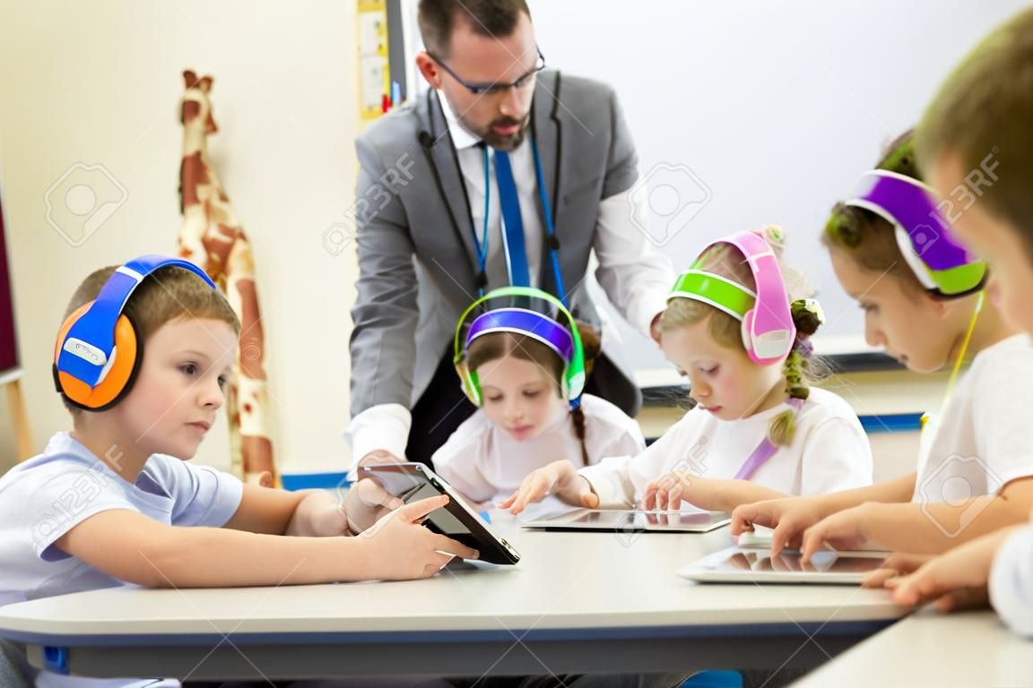 Groupe d'enfants portant des casques sans fil coloré tout en travaillant sur des tablettes numériques, l'enseignant peut être vu superviser les élèves dans la classe