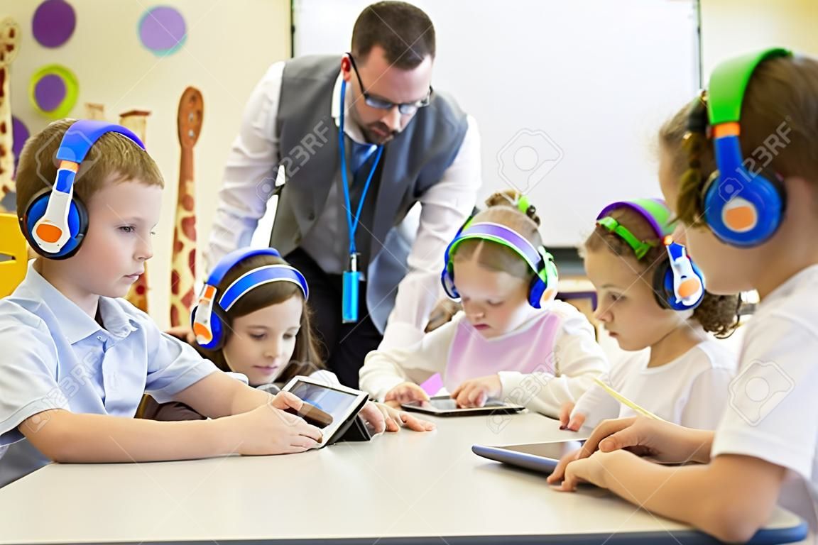 Groupe d'enfants portant des casques sans fil coloré tout en travaillant sur des tablettes numériques, l'enseignant peut être vu superviser les élèves dans la classe