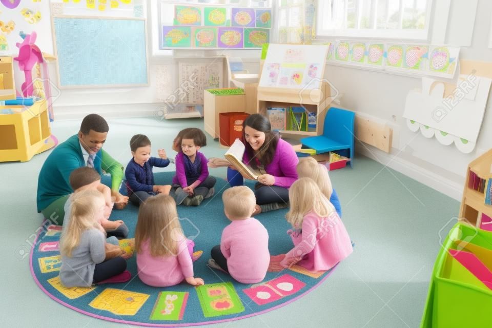 Grupo de crianças do berçário sentado no chão em sua sala de aula com seus professores. A professora fêmea está lendo de um livro.