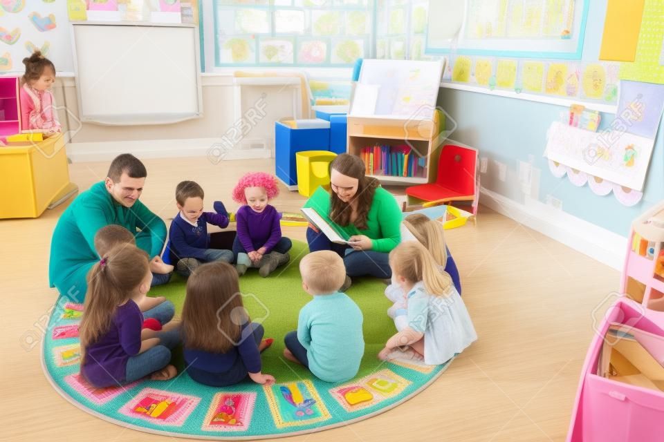 Grupa dzieci przedszkolnych siedzą na podłodze w swojej klasie ze swoimi nauczycielami. Samica nauczyciel czyta z książki.