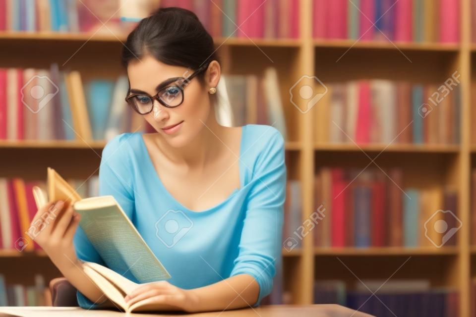 Retrato da mulher jovem bonita, estudante, leitor, senhora, amante da literatura, usando óculos da moda e blusa de algodão que lê um livro que senta-se em uma mesa de madeira na biblioteca, loja de livros.