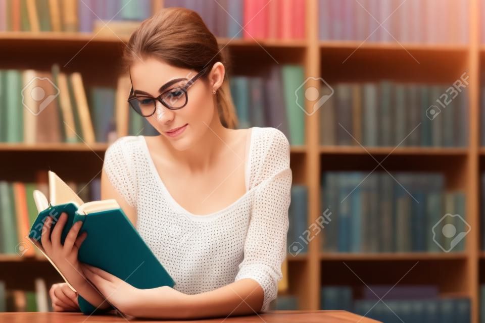 Retrato da mulher jovem bonita, estudante, leitor, senhora, amante da literatura, usando óculos da moda e blusa de algodão que lê um livro que senta-se em uma mesa de madeira na biblioteca, loja de livros.