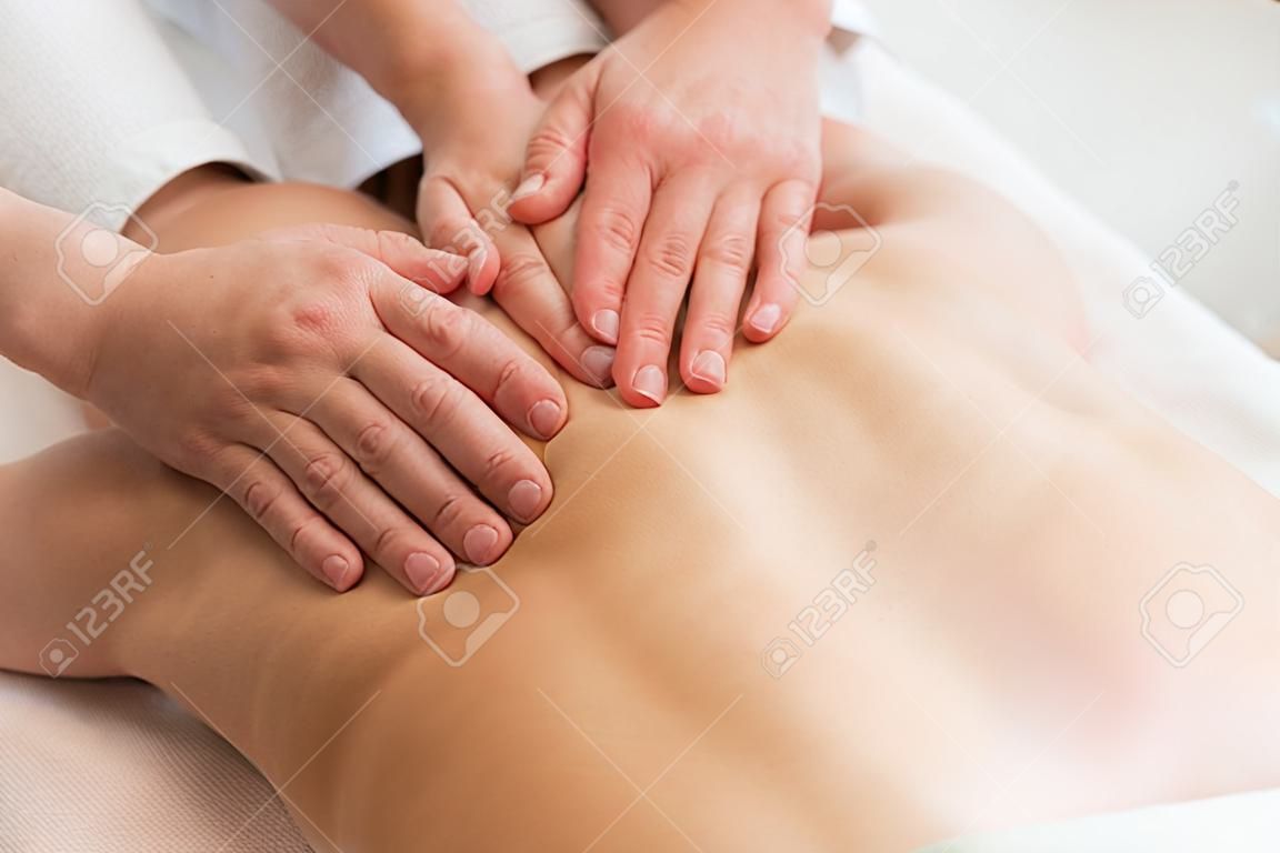 Close-up top view of male masseur massaging lower back of young woman lying on massage table at light spa salon. Experienced chiropractor performs wellness treatments for lady with back pain.
