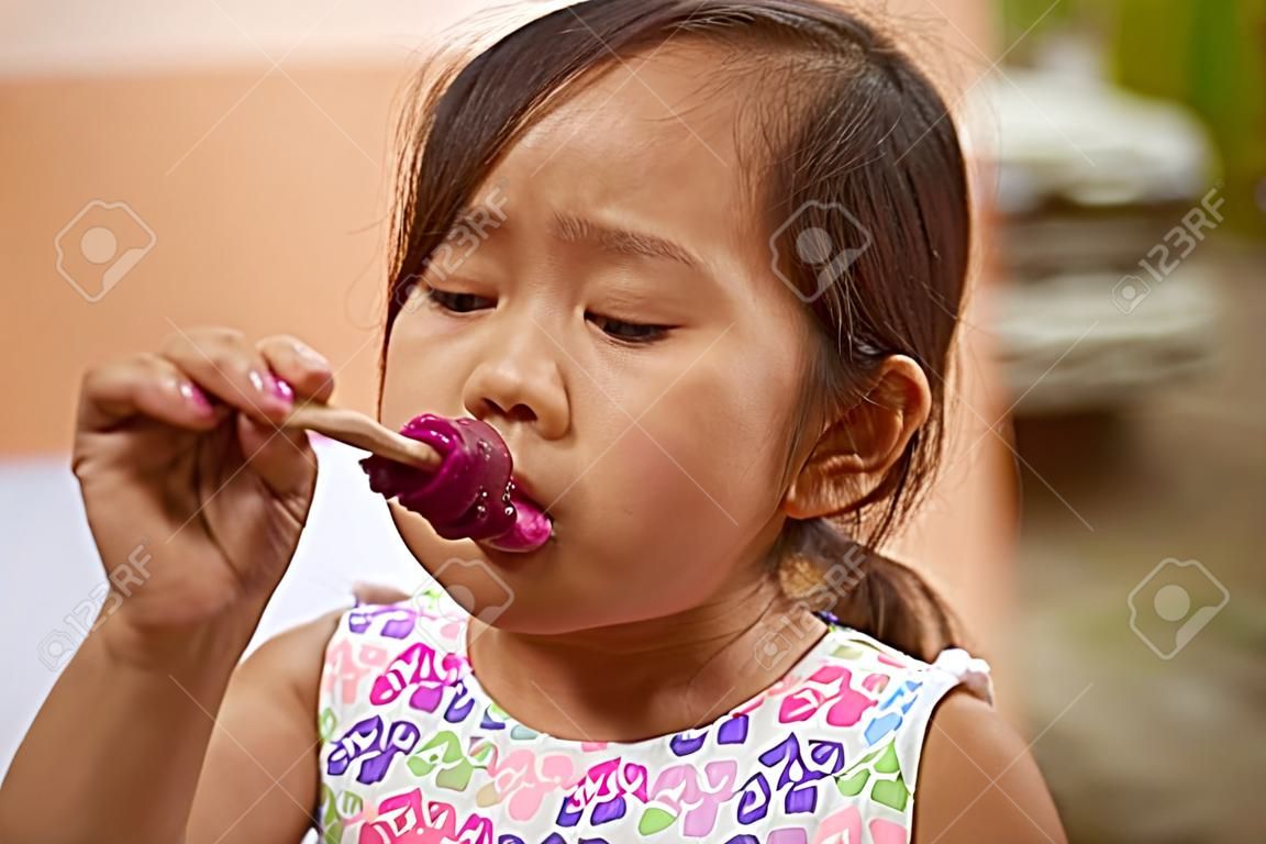 La piccola ragazza asiatica succhia il gelato sul bastone di legno