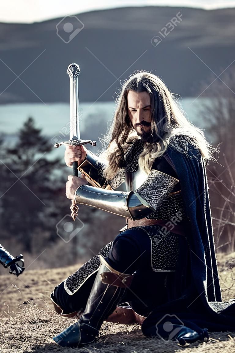 Caballero de pelo largo con espada de dos manos. Fondo rural medieval.