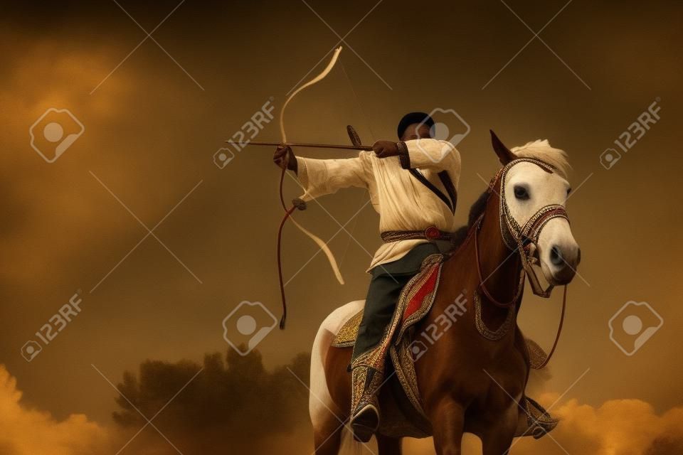 Mężczyzna w stroju etnicznym jedzie na koniu i celuje z łuku.