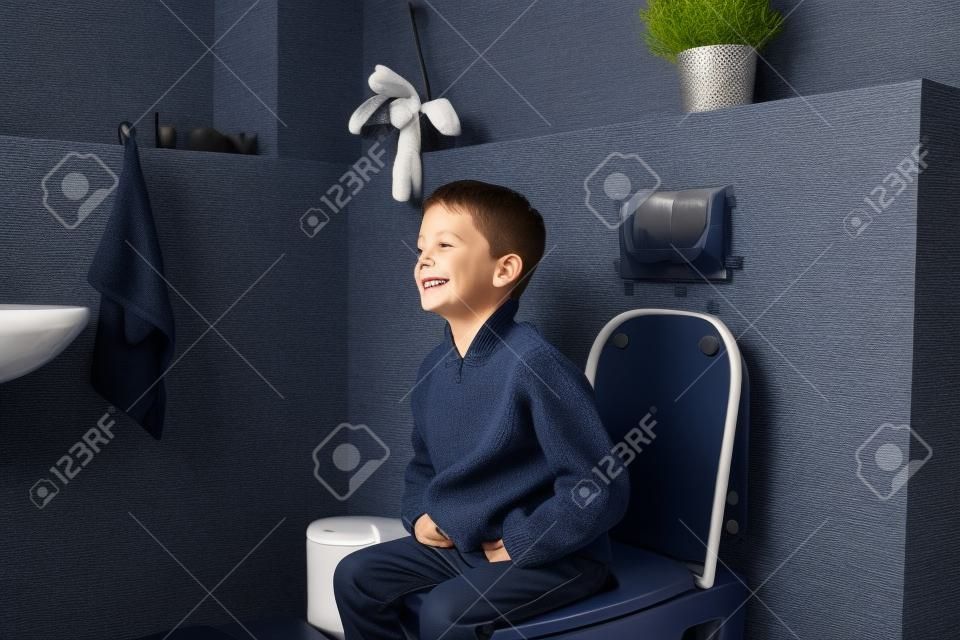 Happy smiling 6 year old boy sitting on the toilet. Dressed in a dark navy blue woolen sweater