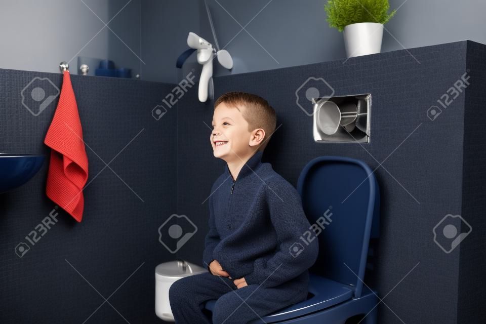 Happy smiling 6 year old boy sitting on the toilet. Dressed in a dark navy blue woolen sweater