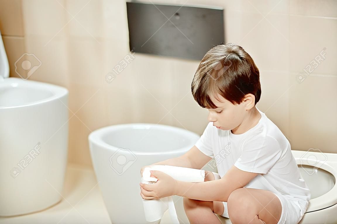 Małe 7 lat chłopiec siedzi na toalecie. Gospodarstwa biały papier toaletowy
