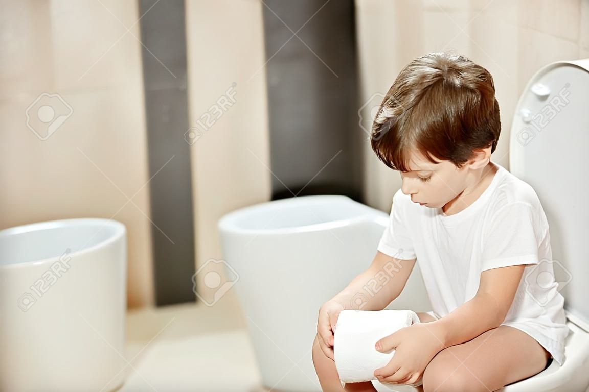 Małe 7 lat chłopiec siedzi na toalecie. Gospodarstwa biały papier toaletowy