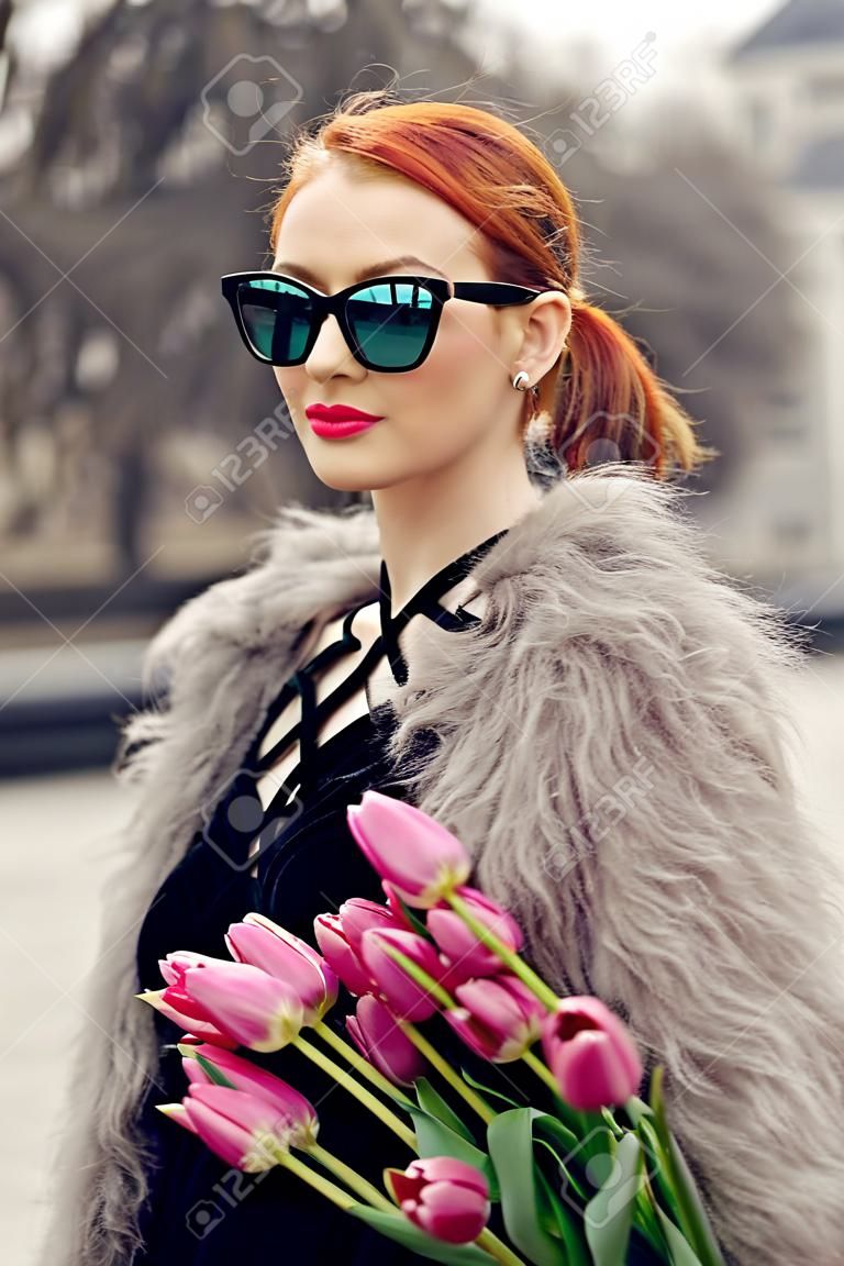 Stijlvol rood haar meisje met paardenstaart in korte zwarte jurk, bontjas en zonnebril. Jonge vrouw met roze tulpen