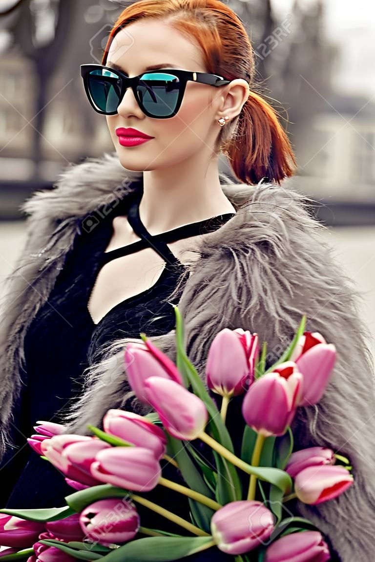 Stylish red hair girl with ponytail wearing short black dress, fur coat and sunglasses. Young woman with pink tulips