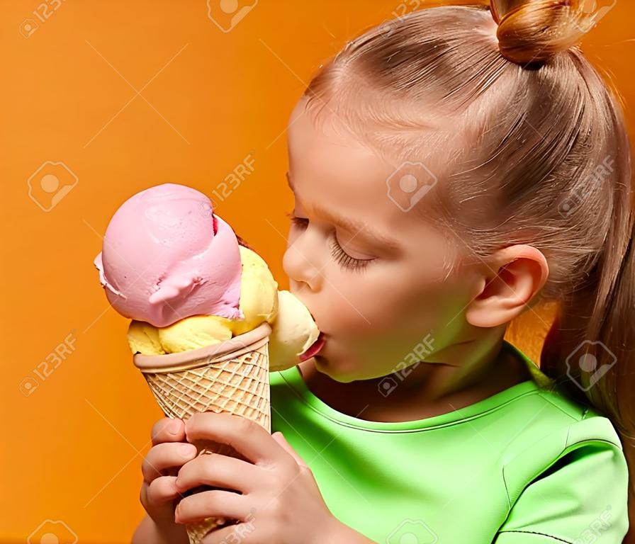 예쁜 아기 소녀 아이가 노란색 배경에 와플 콘에서 바나나와 딸기 아이스크림을 사탕 먹고